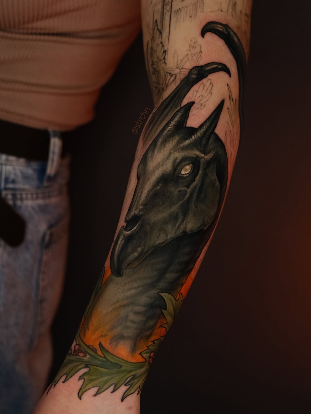 Detailed goat head tattoo on forearm with curved horns, rendered in black and grey with orange and green accents on the lower arm.