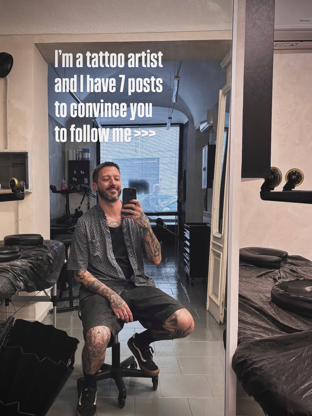 Tattooed man taking mirror selfie in tattoo studio wearing grey cardigan, displaying visible arm and leg tattoos in black ink.