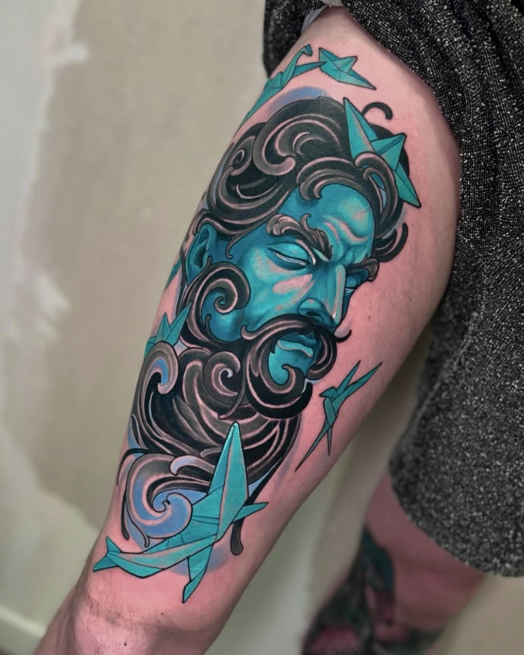 Turquoise bearded male face with ornate dark scrollwork and paper crane designs on upper arm, displayed on light skin tone.