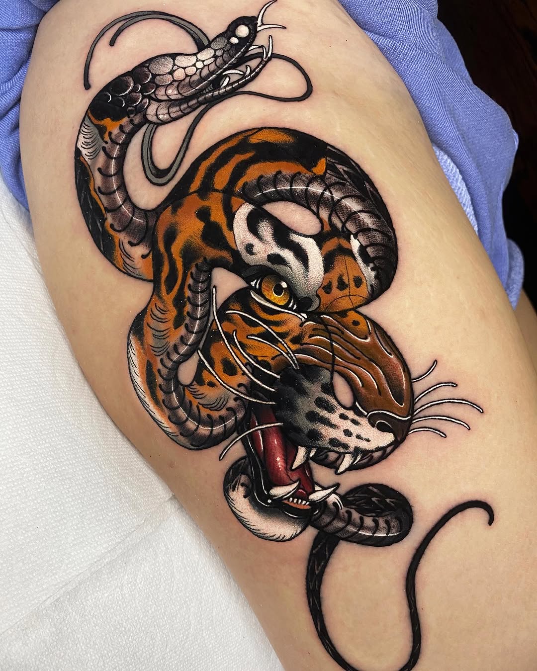 Tiger head with open mouth and intertwined snakes in orange, black, and white ink on upper arm, featuring yellow eyes and detailed scales.