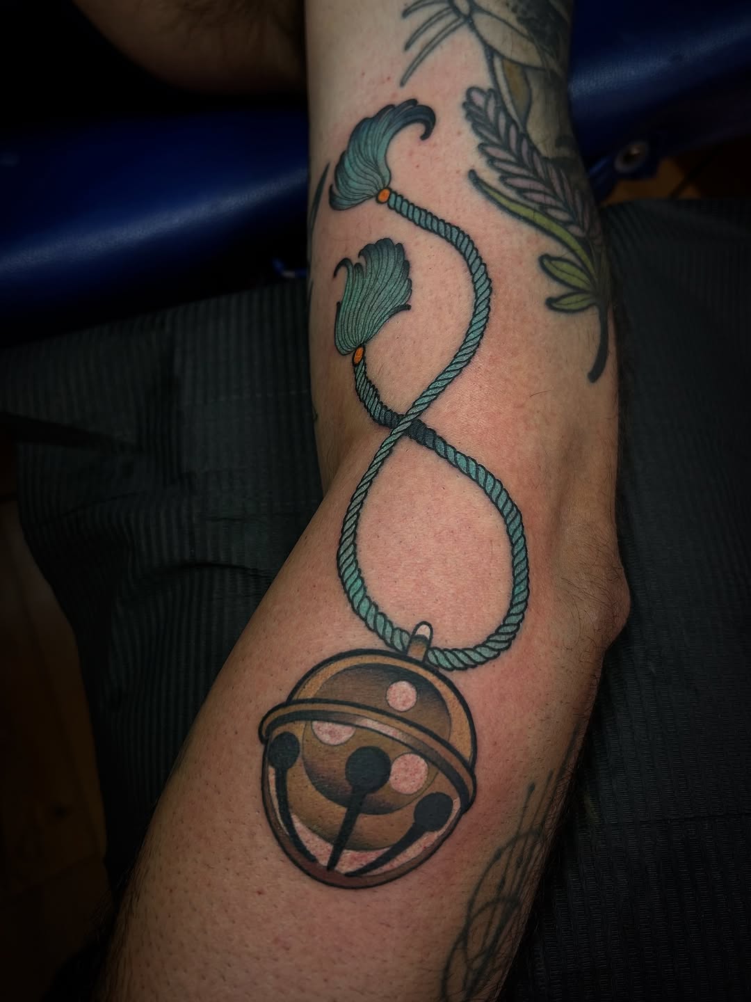 Tattoo on an arm featuring a rope forming an infinity symbol with shell motifs, orange accents, and a circular ladybug design at the base.