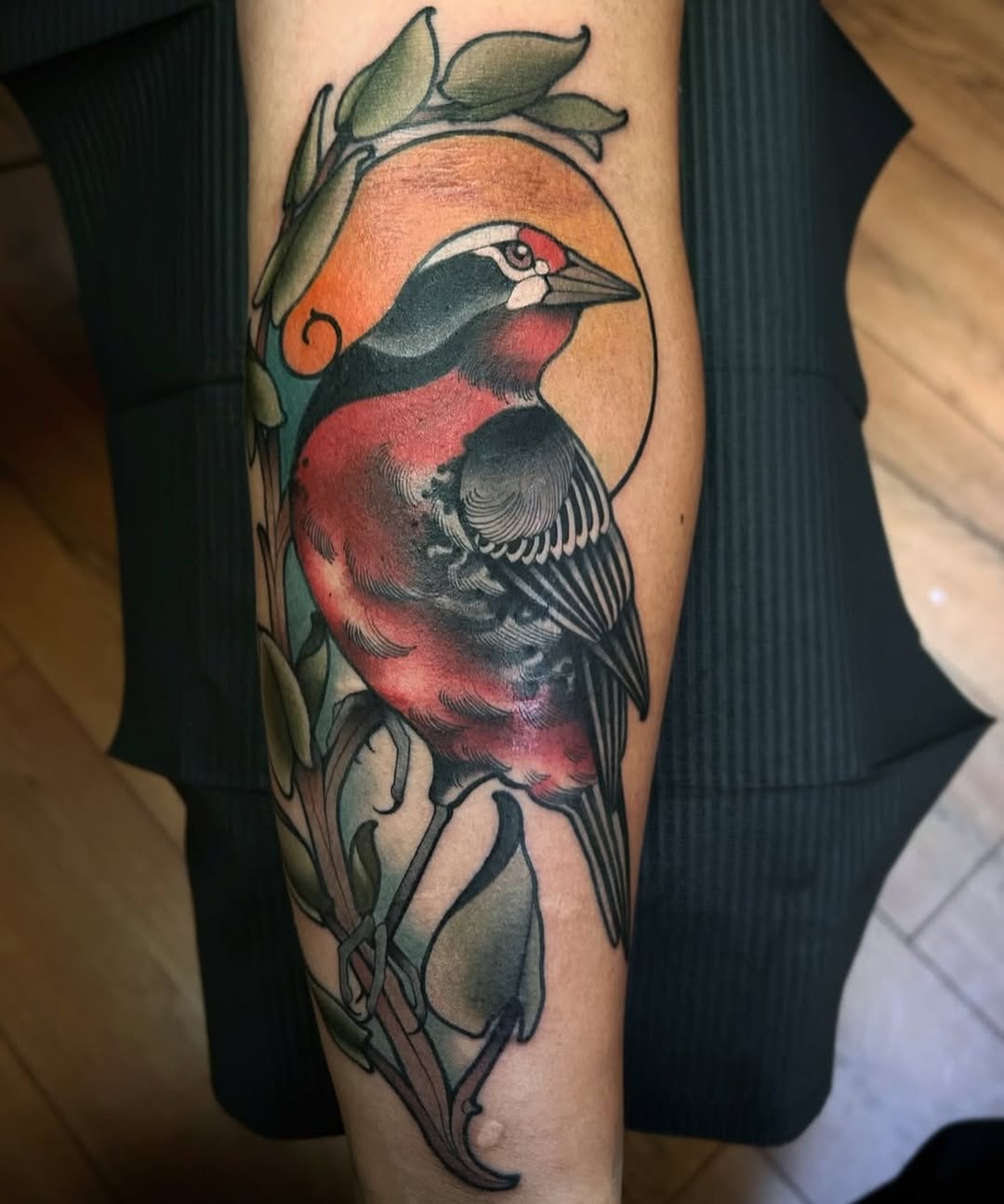 Colorful bird tattoo on forearm featuring red, black, and orange plumage with green leafy branches above and below, shown on pale skin.