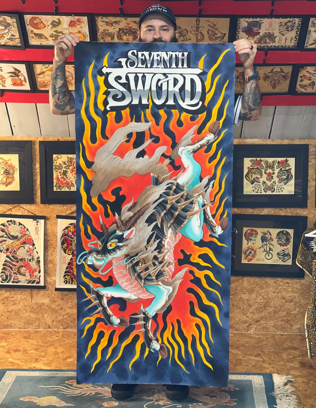 Tattoo artist holding a portfolio print featuring a dragon breathing flames in red, orange, and yellow against a dark blue background with decorative swirls.