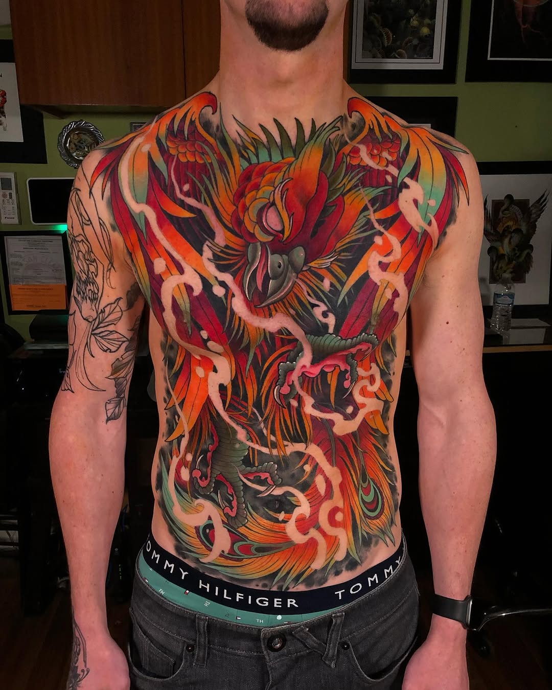 Vibrant chest and torso tattoo featuring red, orange, and black ornamental designs with flowing patterns and bird motifs on a male client.