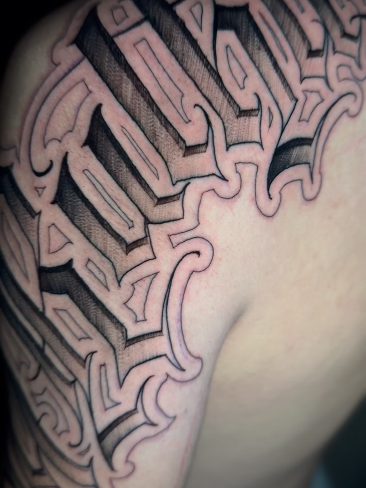 Geometric tribal tattoo with interlocking angular patterns and shading on the upper arm and shoulder in black and grey ink.