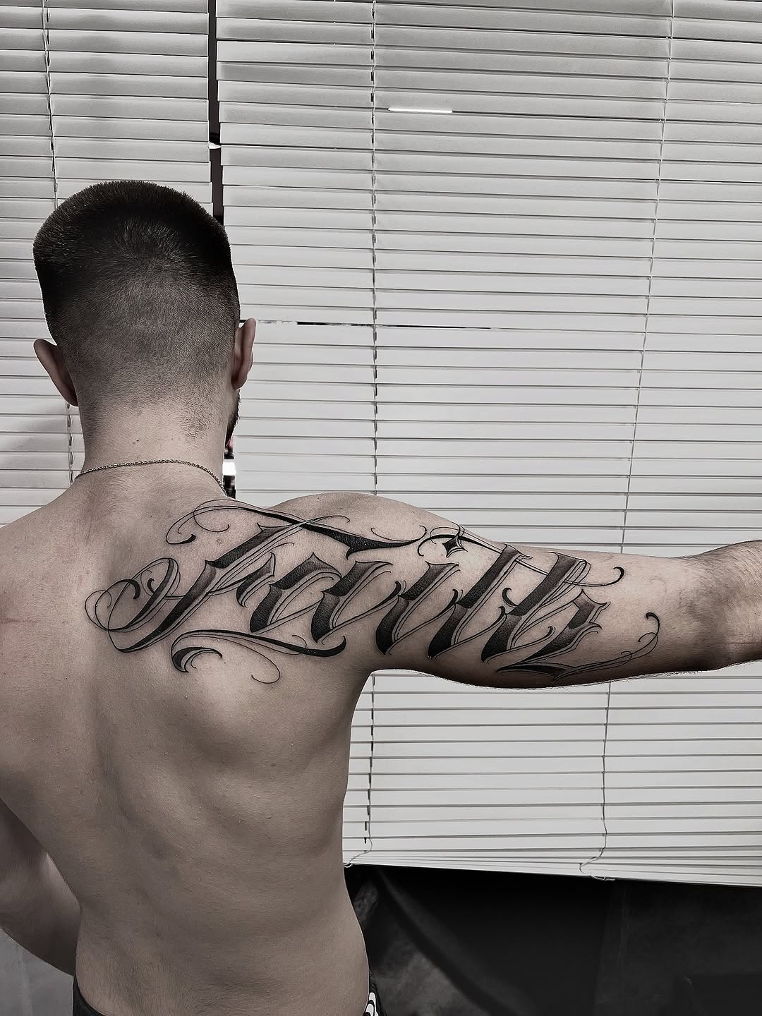 Man displaying a large black script tattoo on his upper back and shoulder with decorative flourishes.