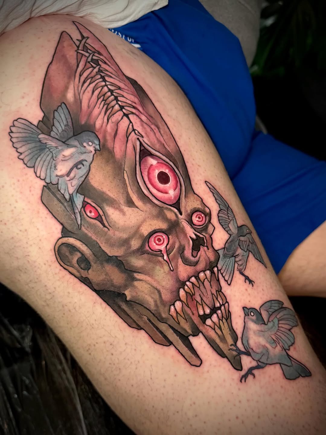 Detailed skull tattoo with fish wings, red eyes, and small birds in flight, positioned on the upper arm in color and black ink.