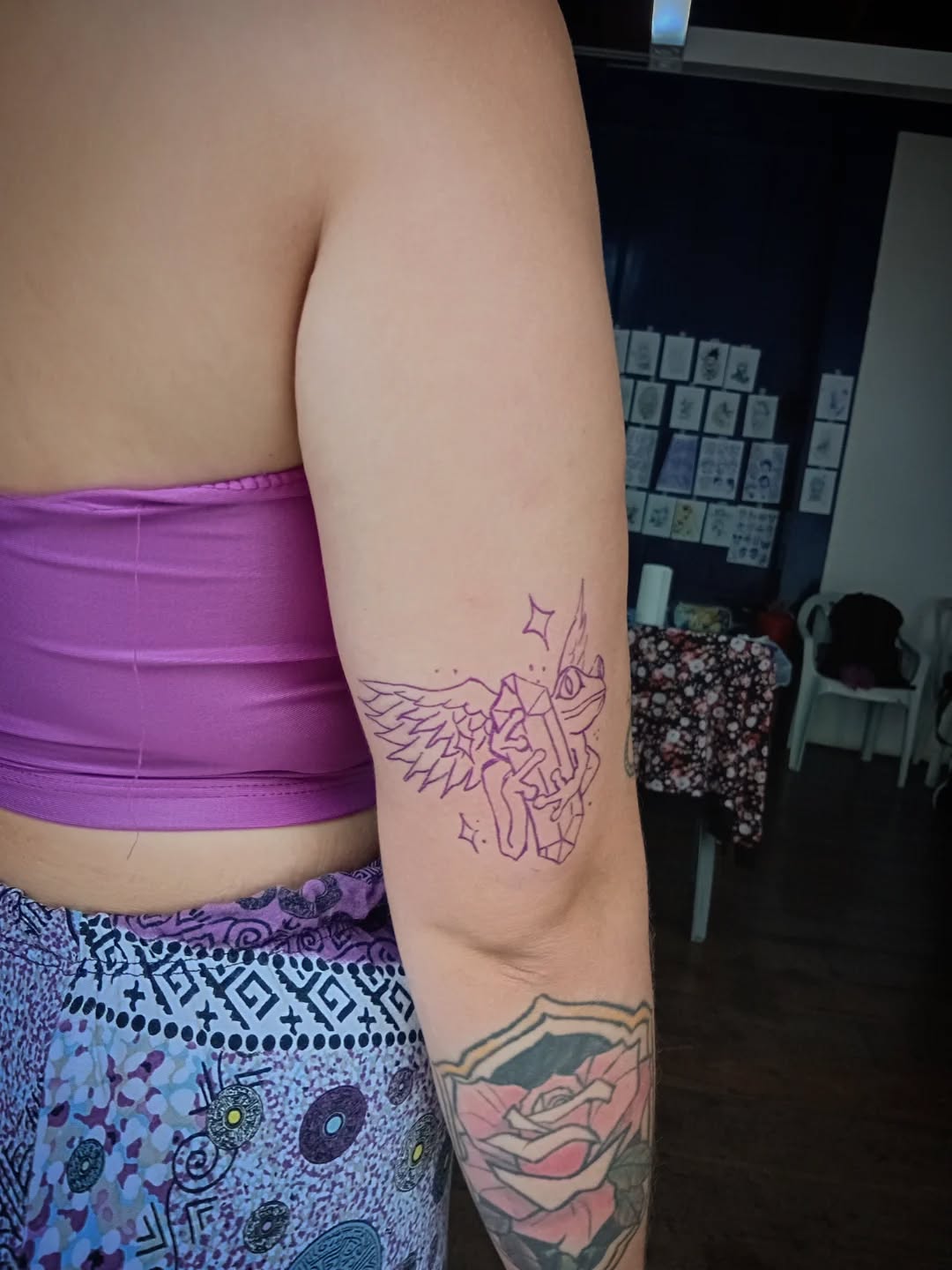 Two tattoos on upper arm: outlined winged figure with stars, and colored hands holding rose design on lower arm.