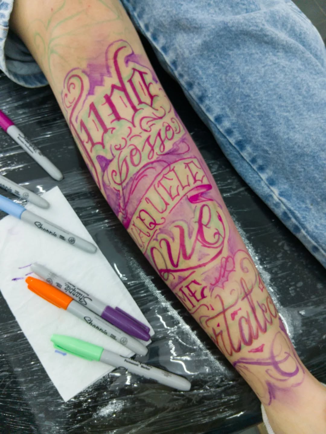 Colorful lettering tattoo on forearm featuring pink and yellow script with decorative swirls, displayed with markers nearby.