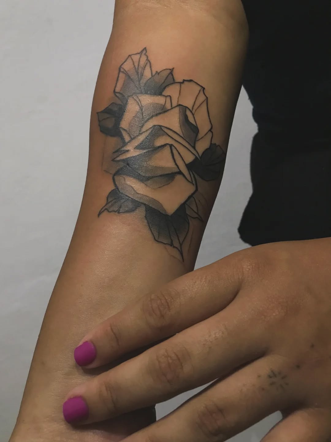 Rose flower tattoo on the upper arm with gray and black shading, shown on a person with purple nail polish.