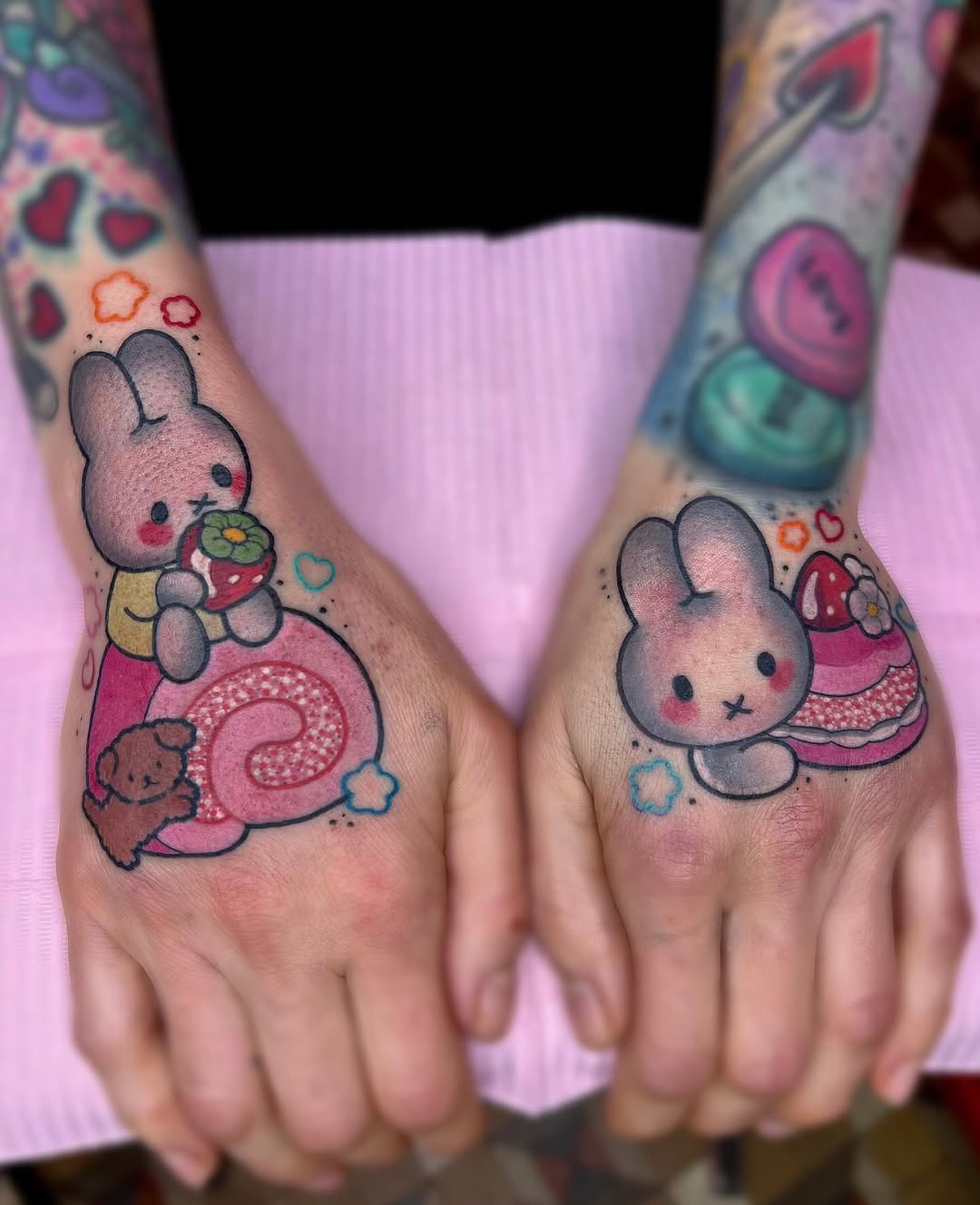 Colorful hand tattoos featuring gray bunnies holding strawberries and pastries surrounded by hearts and flowers in pink, red, and blue tones.