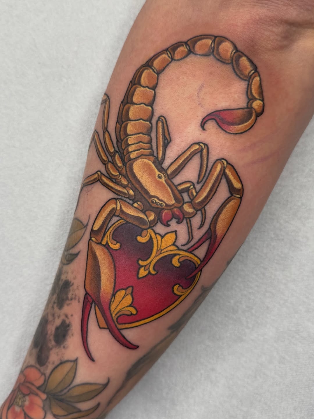 Scorpion tattoo on forearm featuring golden-brown segmented body with red and maroon shield design, ornamental gold flourishes, and decorative elements.