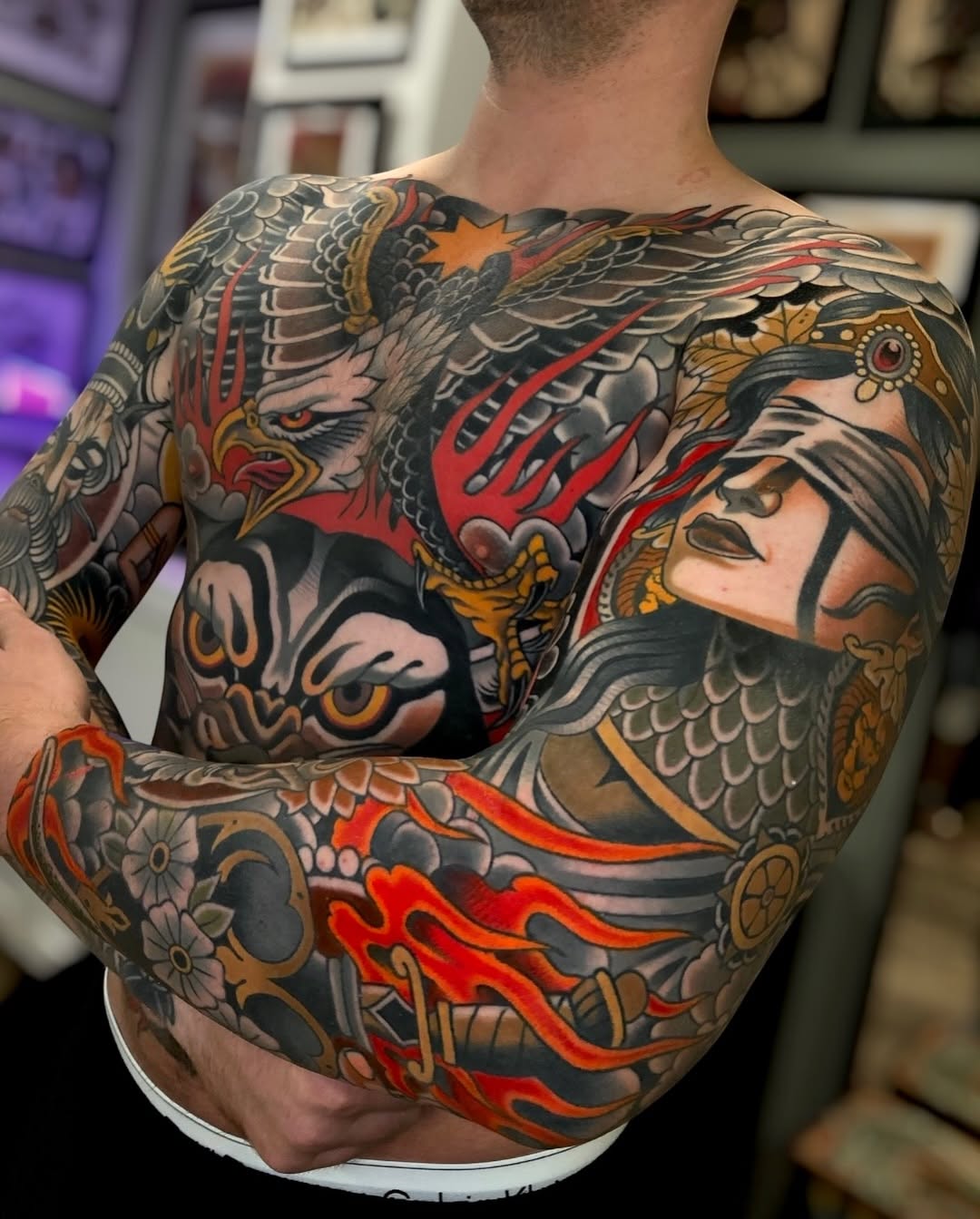 Back tattoo featuring a winged demon face, a bearded warrior portrait, red flames, ornamental patterns, and gold accents in vibrant colors.