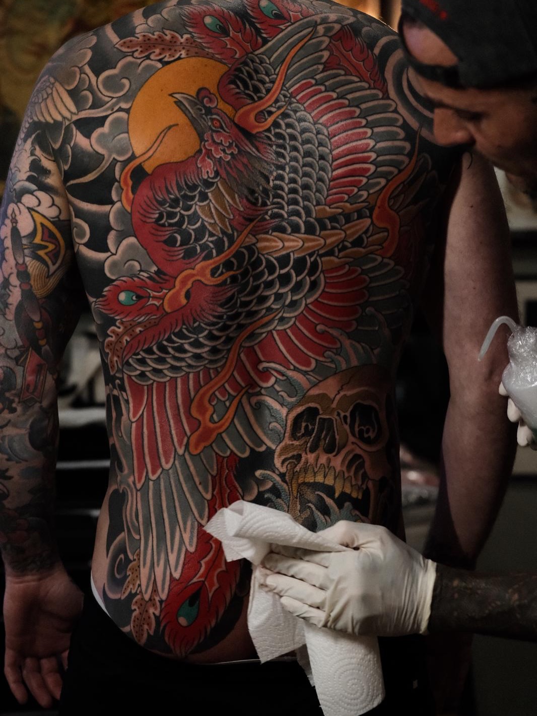 Large phoenix tattoo on chest and shoulder with red, orange, and black feathers, yellow sun, and skull design, being cleaned during session.
