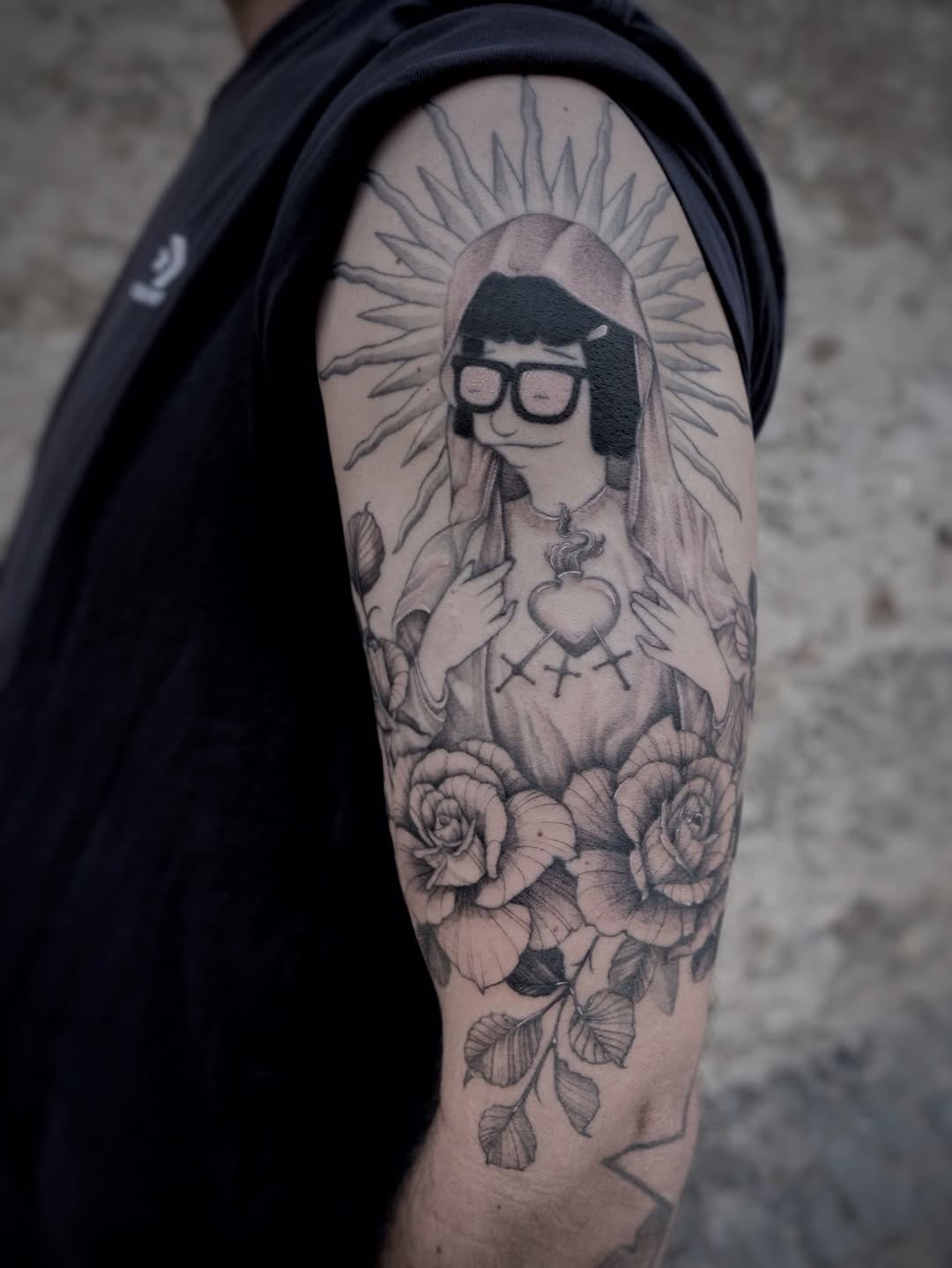 Monochromatic upper arm tattoo featuring a figure with glasses and long hair holding a heart, surrounded by roses and radiating lines.