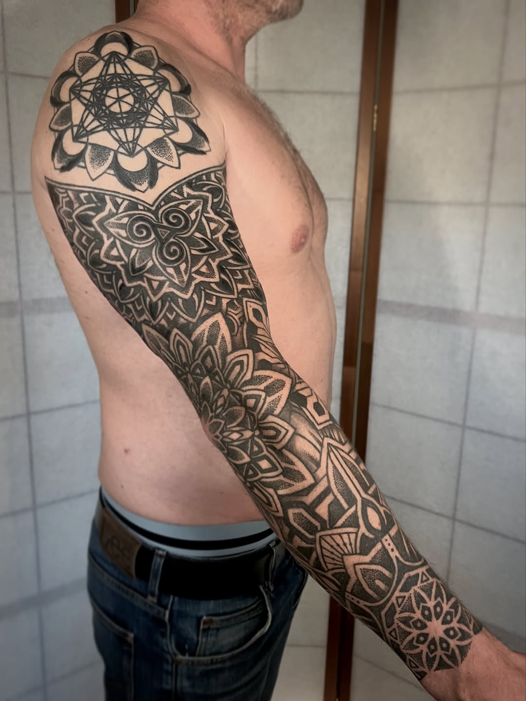 Man displaying a full arm sleeve tattoo featuring geometric mandala and floral patterns in black ink covering shoulder to wrist.