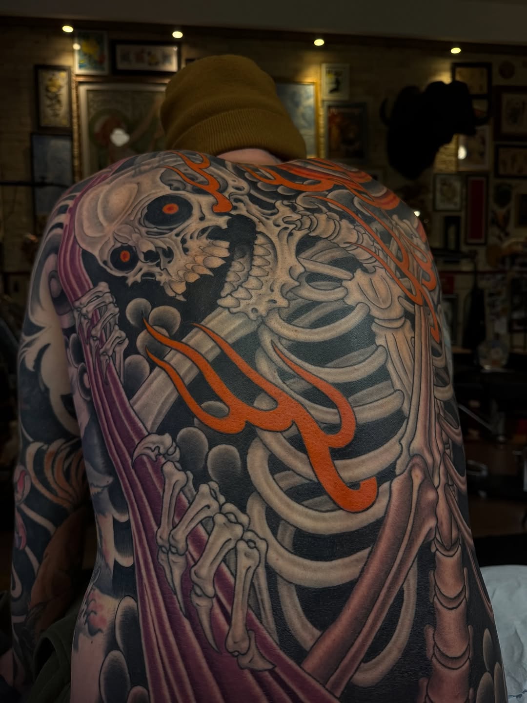 Back and shoulder tattoo featuring a skull with skeletal ribcage, orange flame details, and purple draped fabric elements in multiple colors.