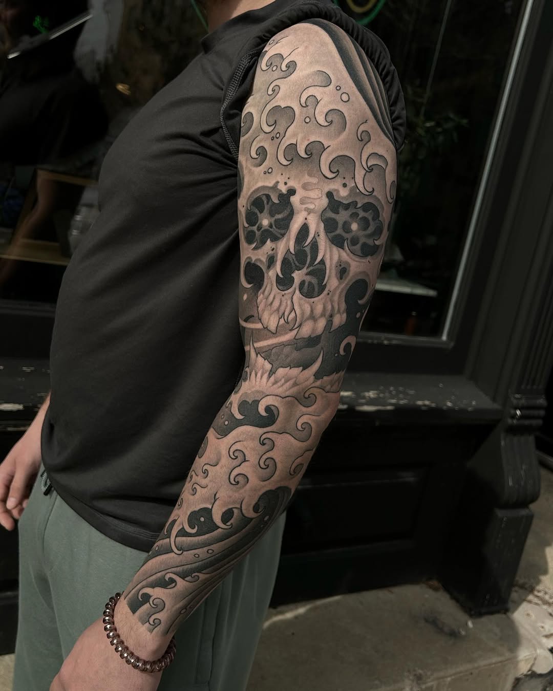 Full arm sleeve tattoo featuring a decorative skull with ornate swirling patterns in gray and black ink on the shoulder and forearm.