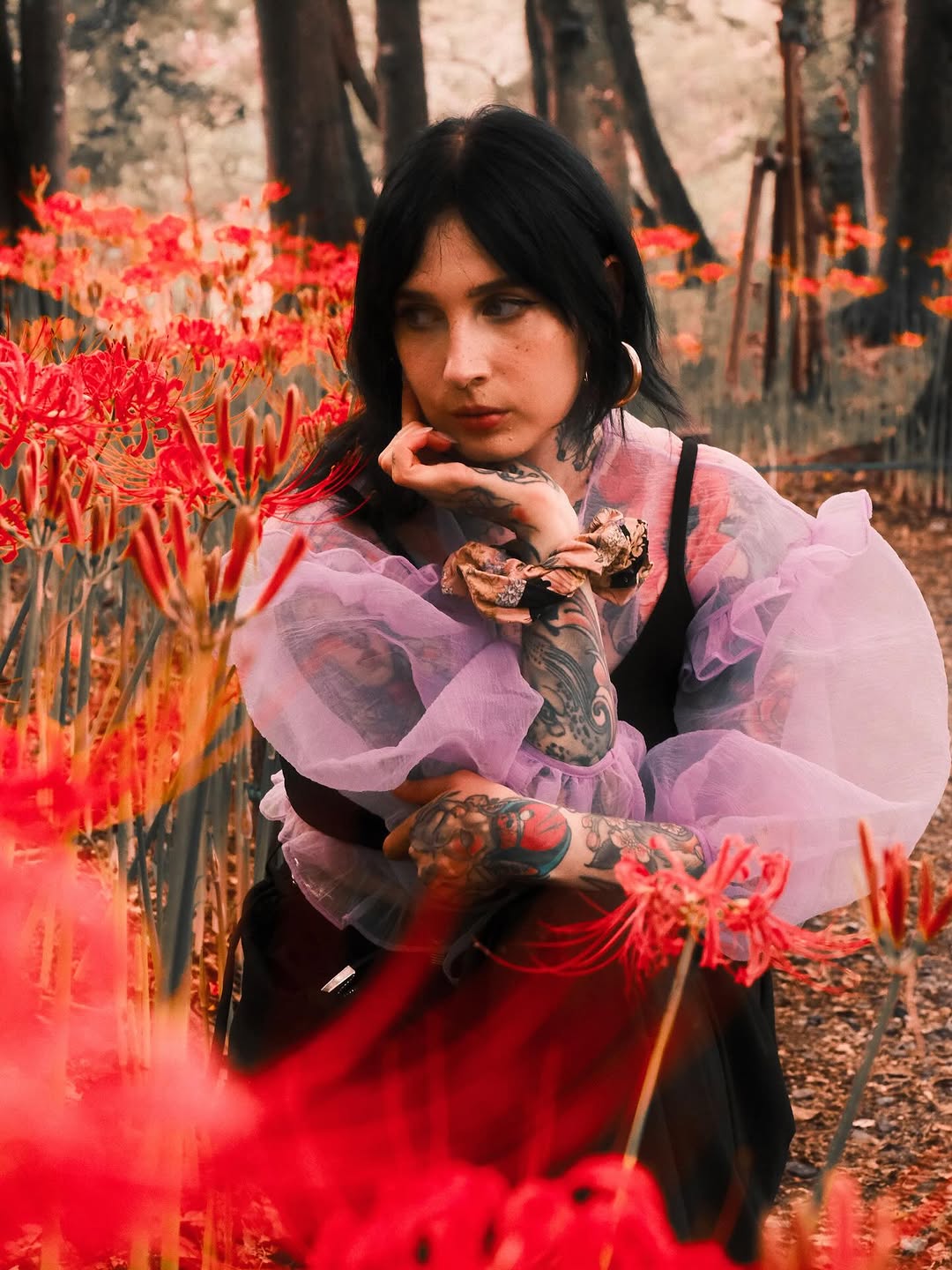 Woman with full-sleeve arm tattoos in color and black ink poses among red spider lily flowers in a forest setting, wearing pink tulle.