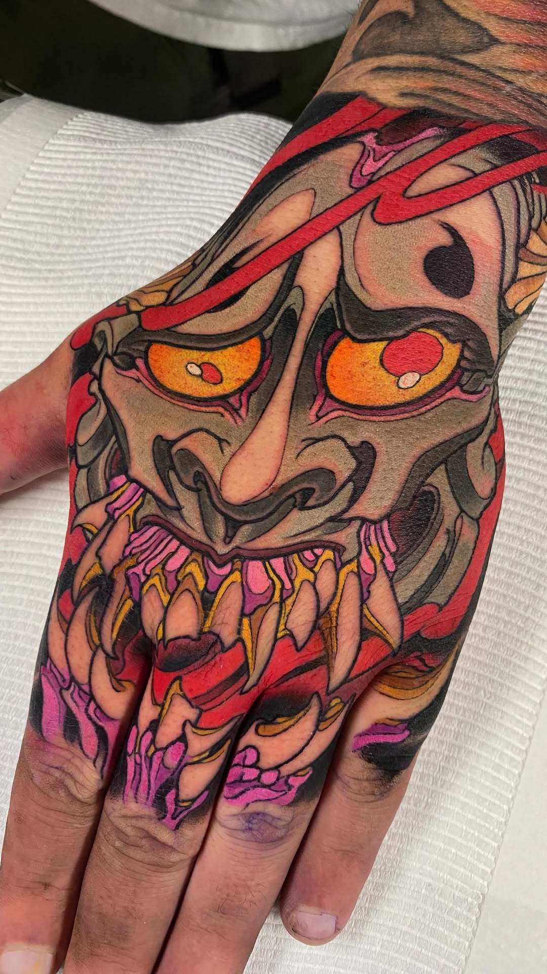 Colorful demon or oni mask tattoo on hand featuring orange eyes, red ribbon, purple and gold teeth, with vibrant red, orange, pink, and black ink.