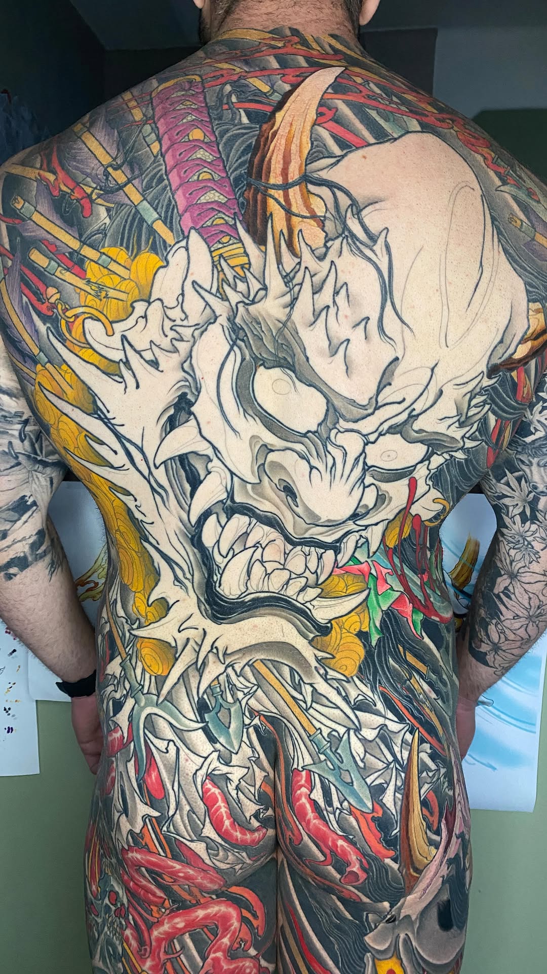 Back tattoo featuring a large skull with anatomical details in cream and black, surrounded by colorful flowers and foliage in red, yellow, and green tones.