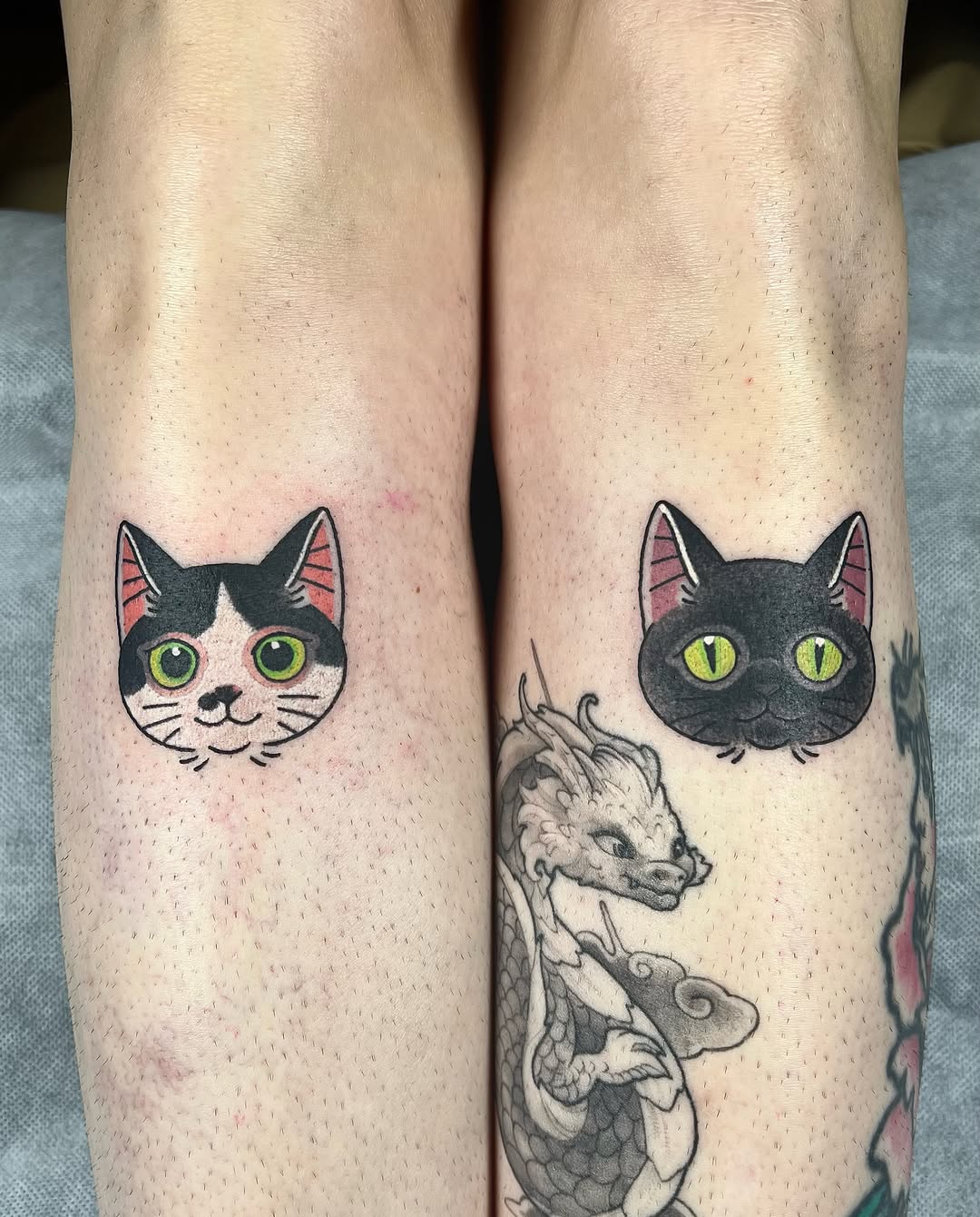 Two cat head tattoos with green eyes and pink inner ears on the calves, with a dragon tattoo visible on the right leg.
