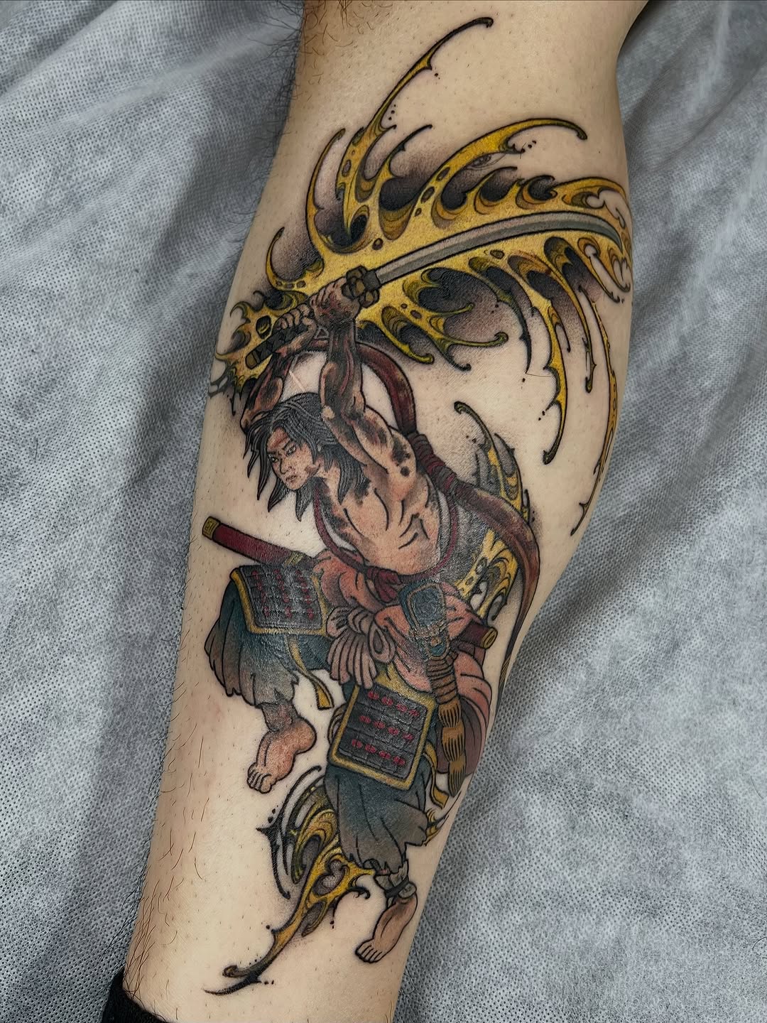 Colorful tattoo on a leg featuring a mythical warrior figure in ornate armor wielding a spear beneath a decorative winged crest in gold and black.