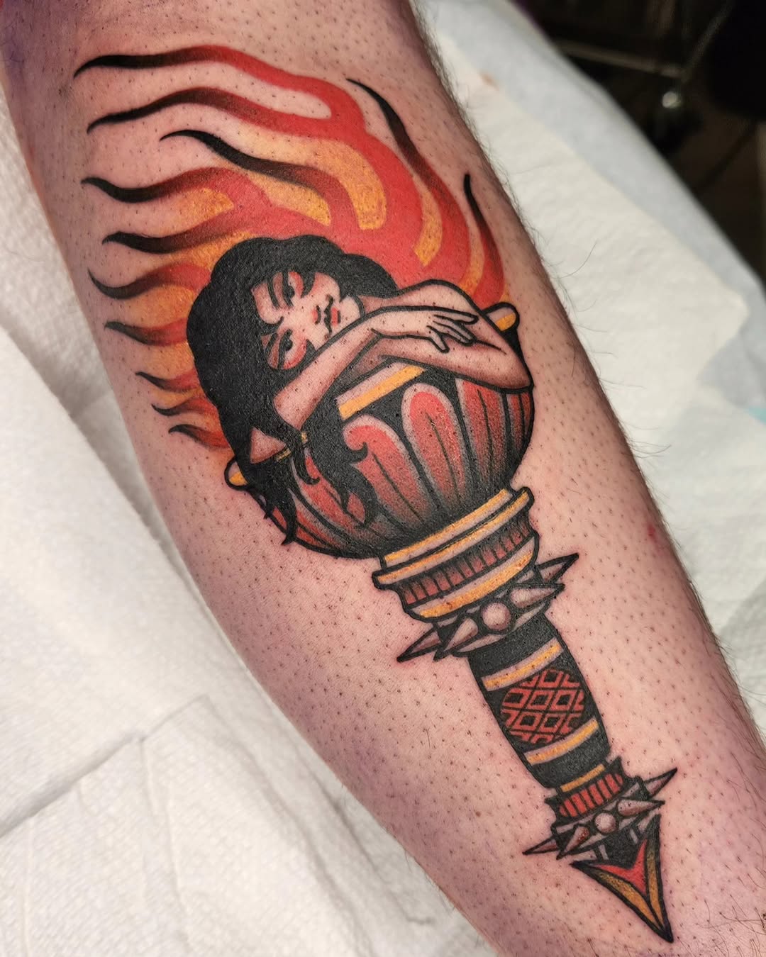 Tattoo on forearm featuring a person's face emerging from a flaming torch with black outlines, red and orange flames, and gold accents on the handle.