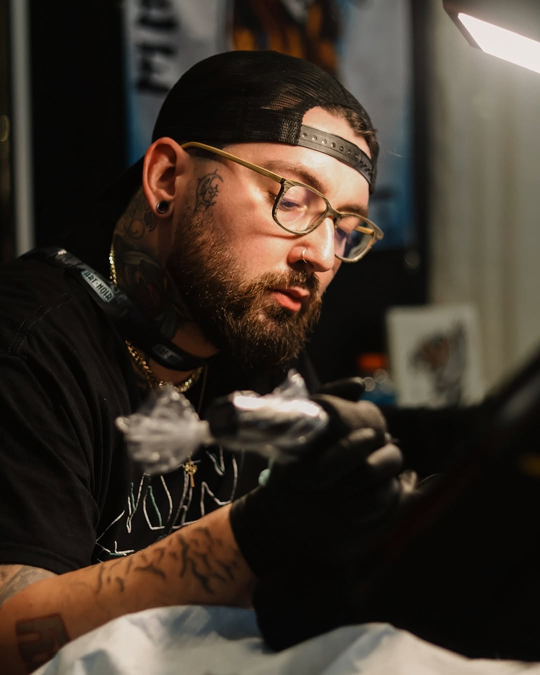 Tattoo artist wearing glasses and headband works on a client's arm with black ink and gloved hands under bright studio lighting.