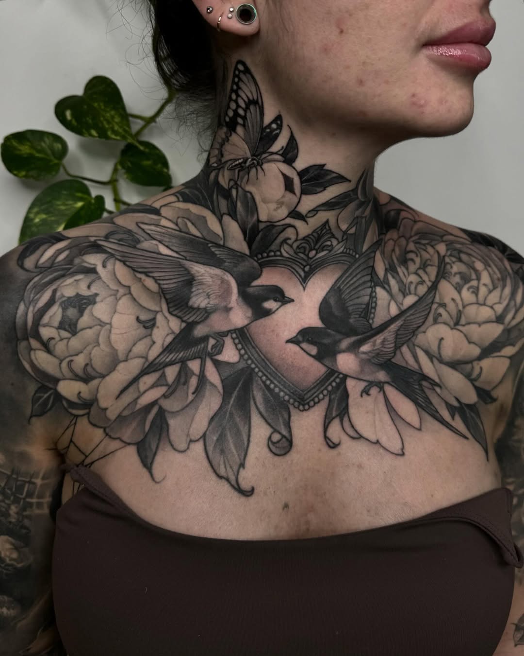 Large chest and shoulder tattoo featuring birds, flowers, and ornamental details in black ink on pale skin, displayed on a female model.