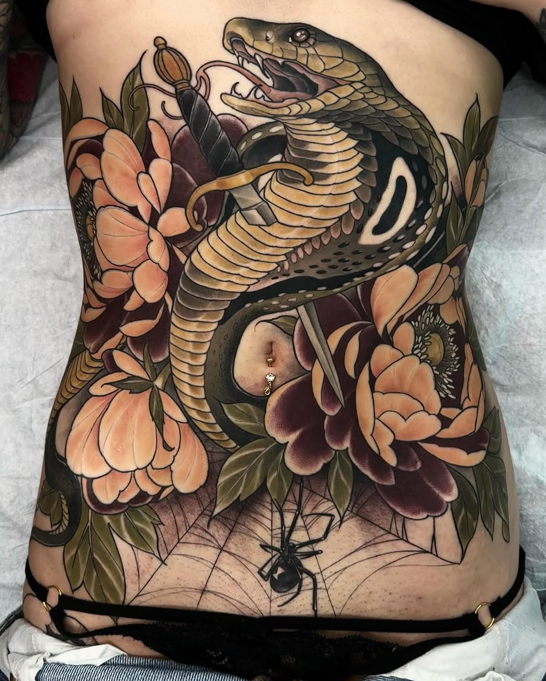 Cobra with mouth open surrounded by peach and burgundy flowers with green leaves and spider web on lower back, tan and gold tones.
