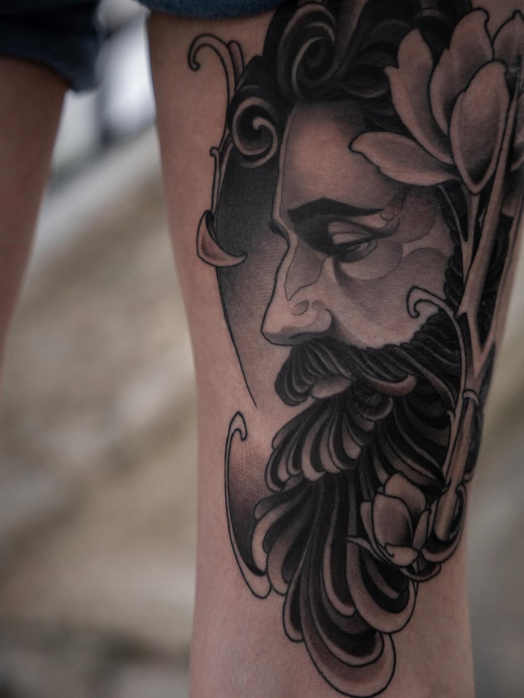 Bearded male face portrait tattoo with decorative scrollwork and floral elements on the leg, rendered in black and gray ink.
