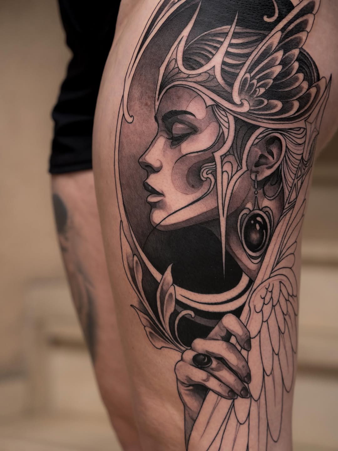 Detailed portrait tattoo on thigh of a winged female figure with ornate headdress, earrings, and decorative linework in black and grey ink.