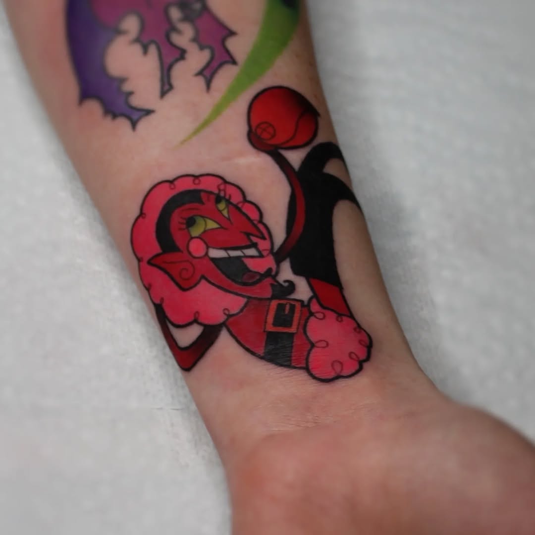 Red and black cartoon character tattoo on forearm featuring a smiling demon-like figure with yellow eyes, horns, and a belt buckle.