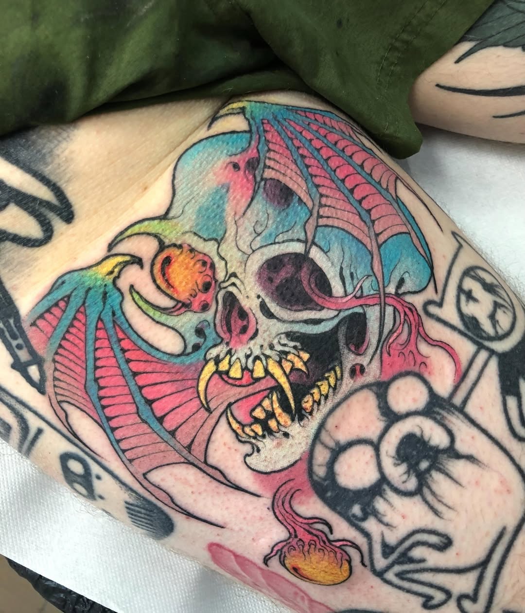 Colorful skull tattoo with bat wings, golden teeth, and jellyfish details on the forearm, featuring turquoise, pink, and orange hues.