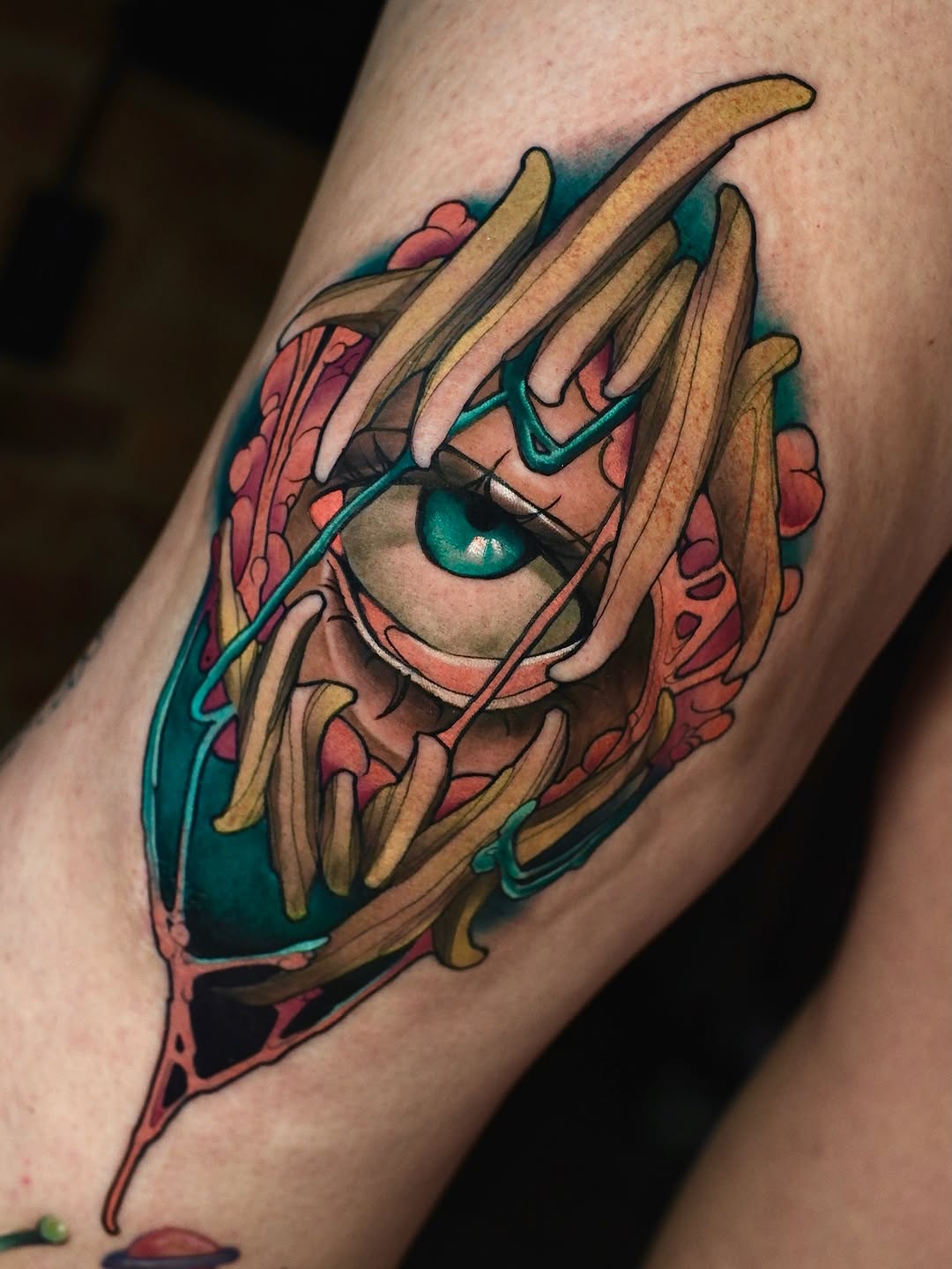 Colorful tattoo on an arm featuring a face with teal eyes and long pointed fingers, surrounded by coral and teal accents.
