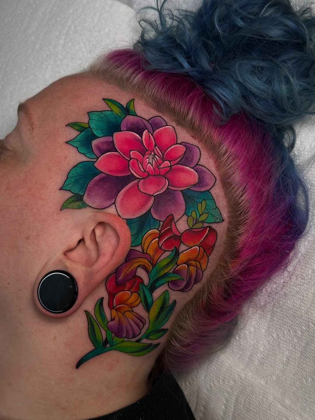 Colorful floral tattoo on the side of the head featuring a pink dahlia with green leaves and orange and red flowers below the ear.