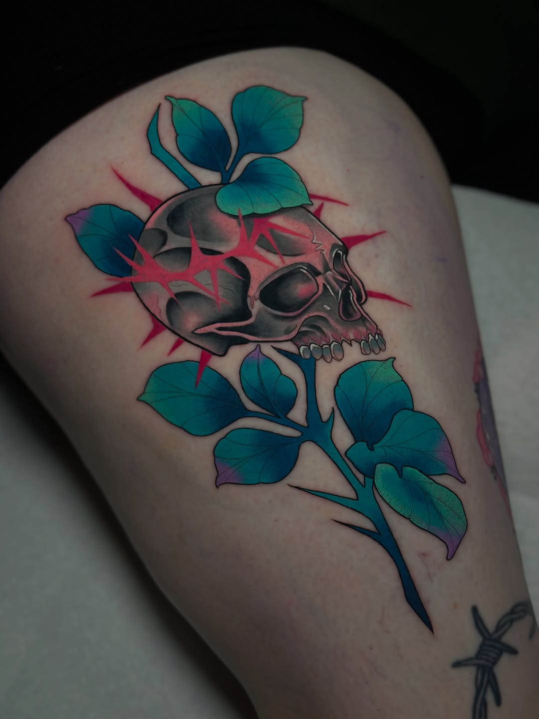 Skull with teal leaves and pink lightning bolts tattooed on upper thigh, featuring detailed shading and color gradients.