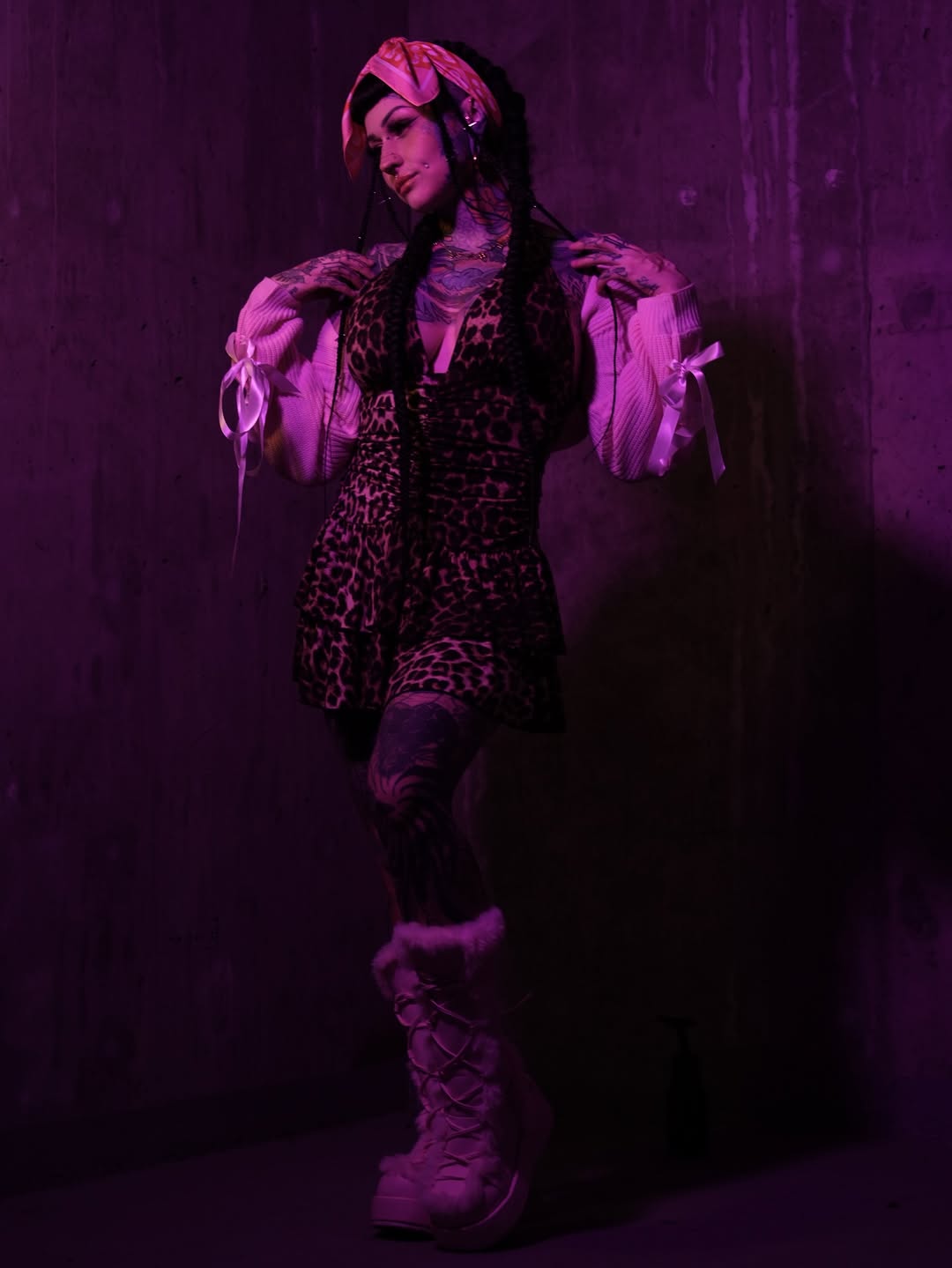 Person wearing a leopard print bodysuit with pink ribboned shoulders and a pink headwrap, photographed against a purple background under pink lighting.