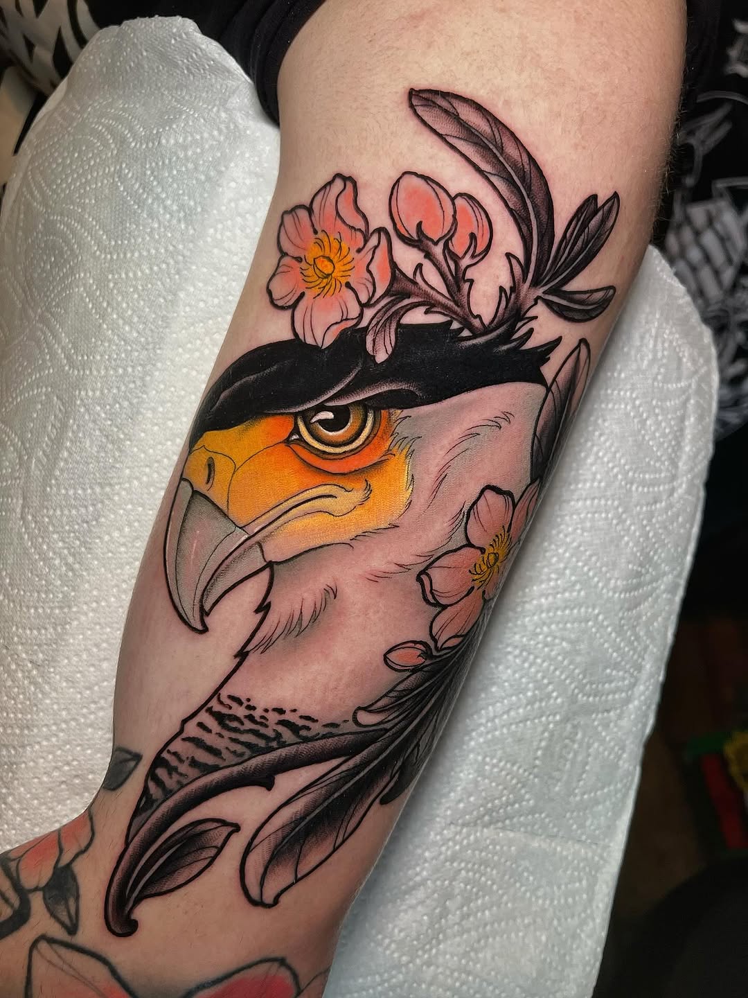Colorful bird head with golden eye and black eye mask adorned with pink flowers on the upper arm, displayed on pale skin.