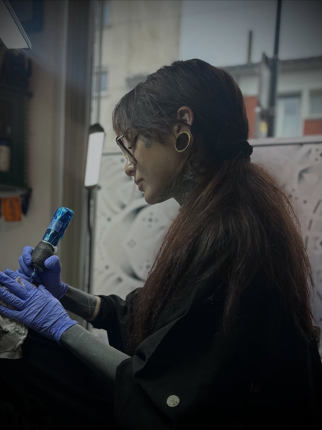 Tattoo artist wearing blue gloves holds a client's arm displaying a small dark tattoo on the inner forearm near the wrist.