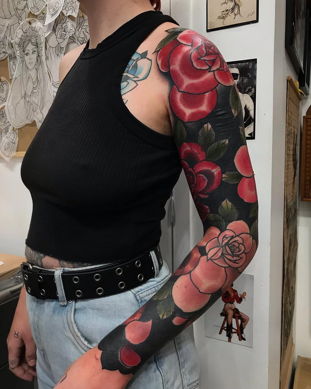 Full-sleeve arm tattoo featuring red and pink roses with green leaves on black background, displayed on person wearing black sleeveless top.