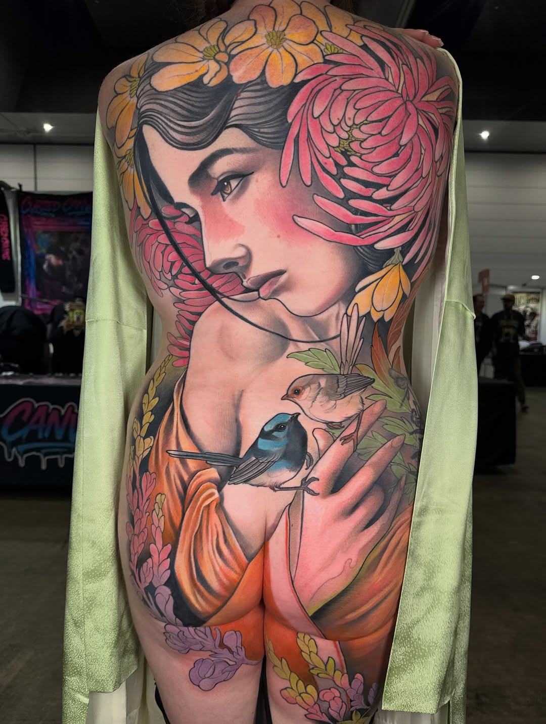 Large-scale back tattoo featuring a woman's profile surrounded by colorful flowers and a small bird, predominantly in orange, pink, and yellow tones.