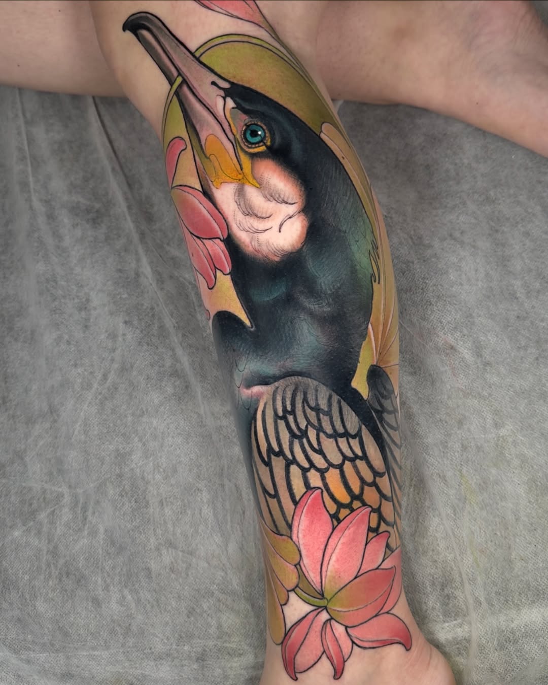 Colorful toucan tattoo on forearm featuring the bird's head and wings with pink lotus flowers and yellow and green accents.