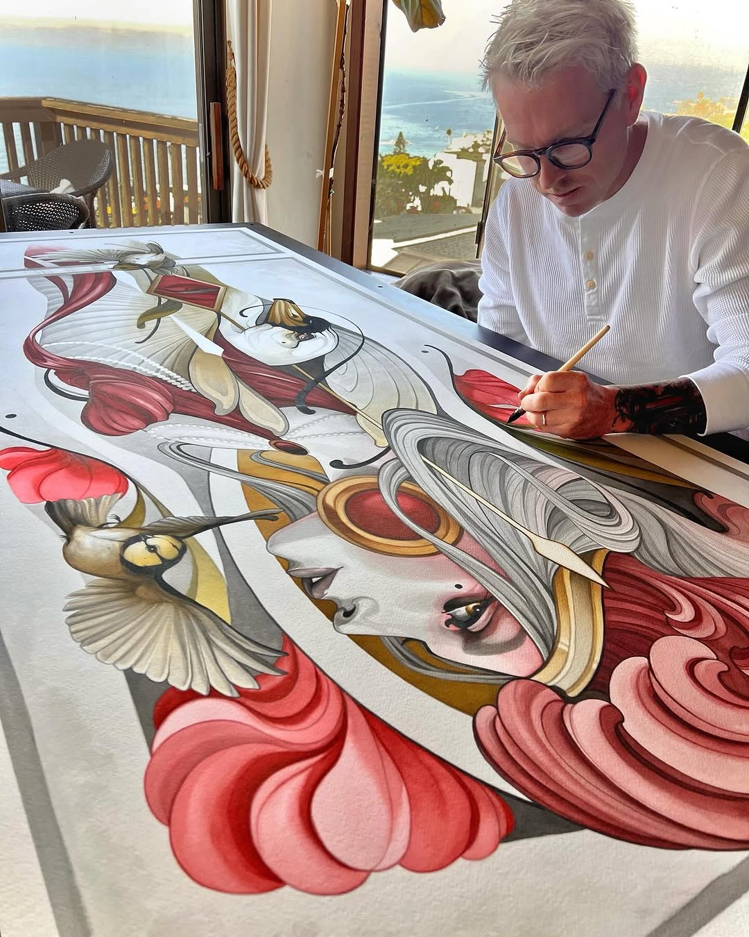 Man with glasses painting an intricate design featuring ornate faces, flowing red and pink petals, gold accents, and detailed linework on large white paper.