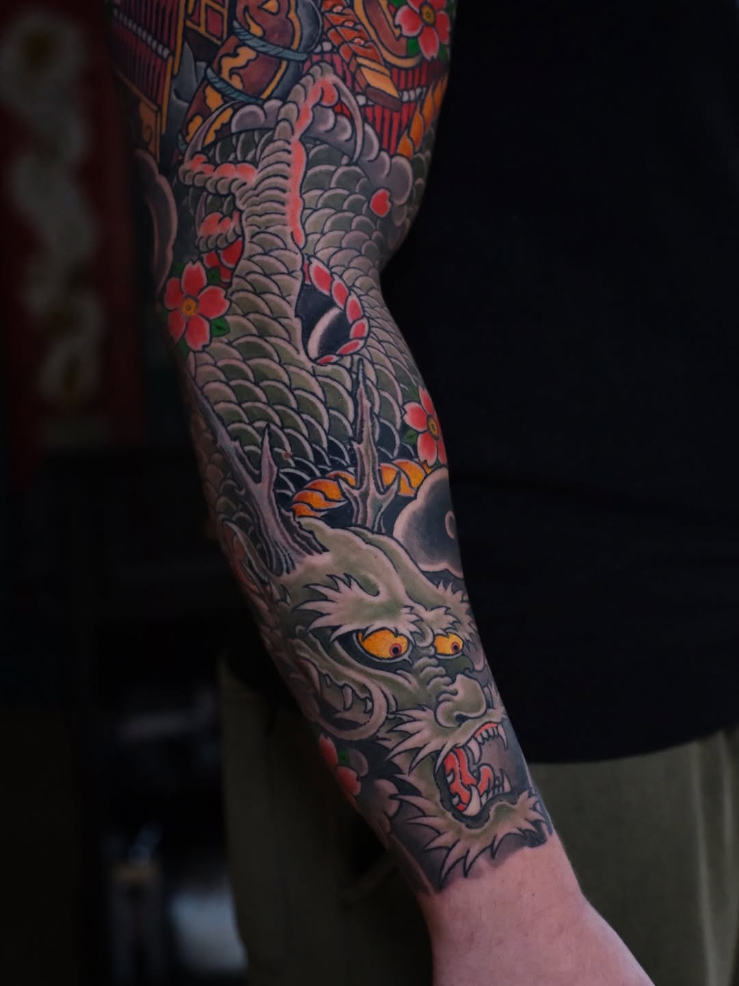 Colorful full-arm sleeve tattoo featuring a dragon with scales, flowers, and a demon mask with orange eyes on the forearm.