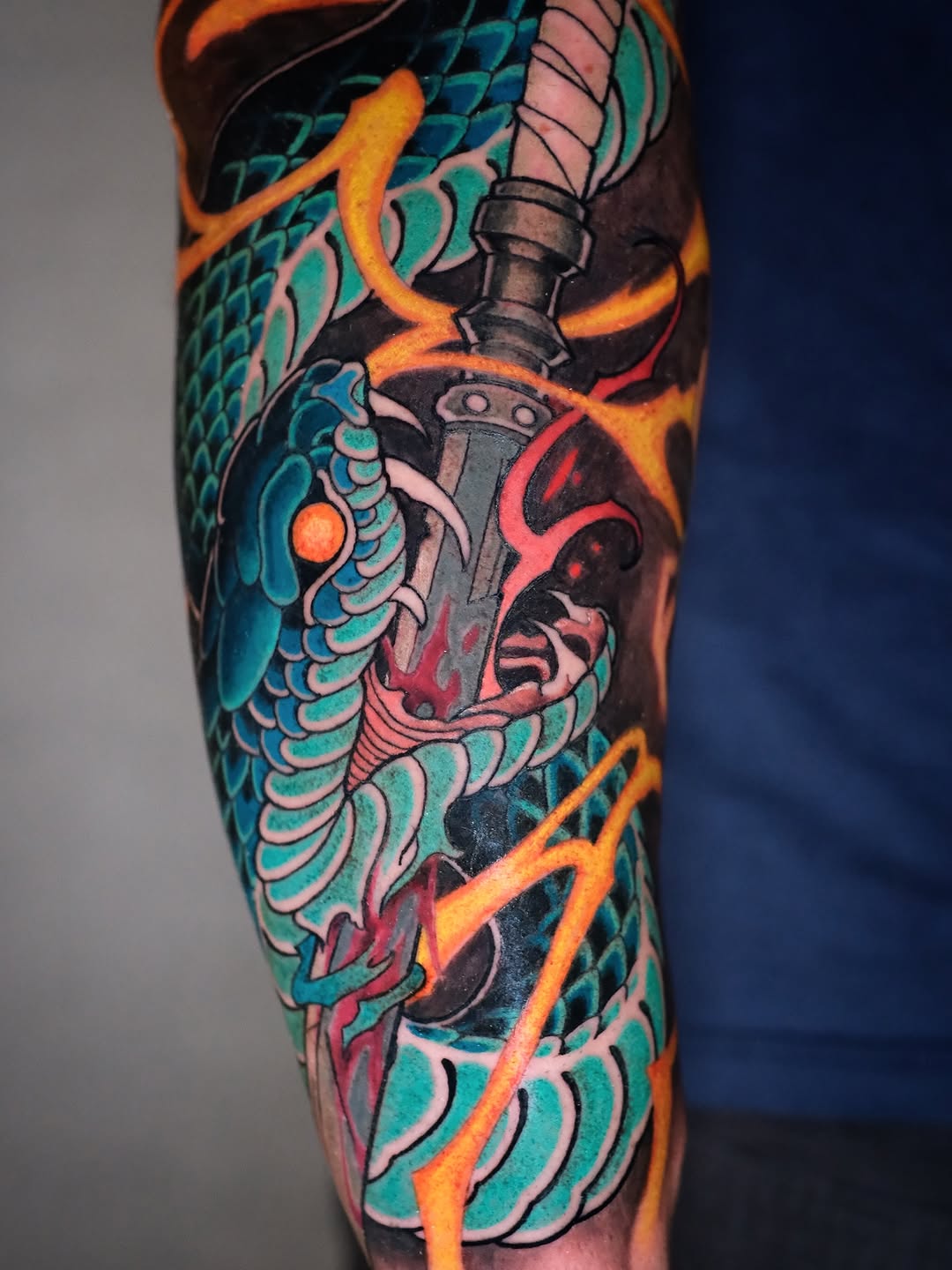 Colorful sleeve tattoo featuring a dragon with turquoise scales, a demon face in red and pink, a dagger, and orange flame elements on the upper arm.