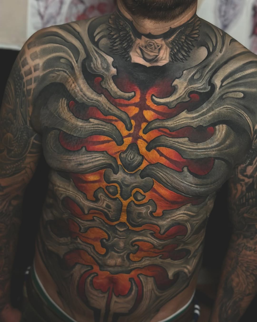 Detailed chest tattoo featuring ornamental grey scrollwork framing a central glowing orange and red flame design with a rose and winged motif at the neckline.