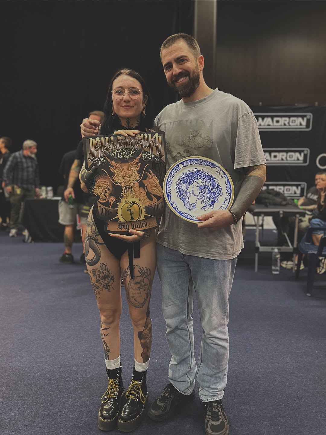 Two people display extensive leg and body tattoos featuring animals, skulls, and ornamental designs in black and colored ink at an indoor event.