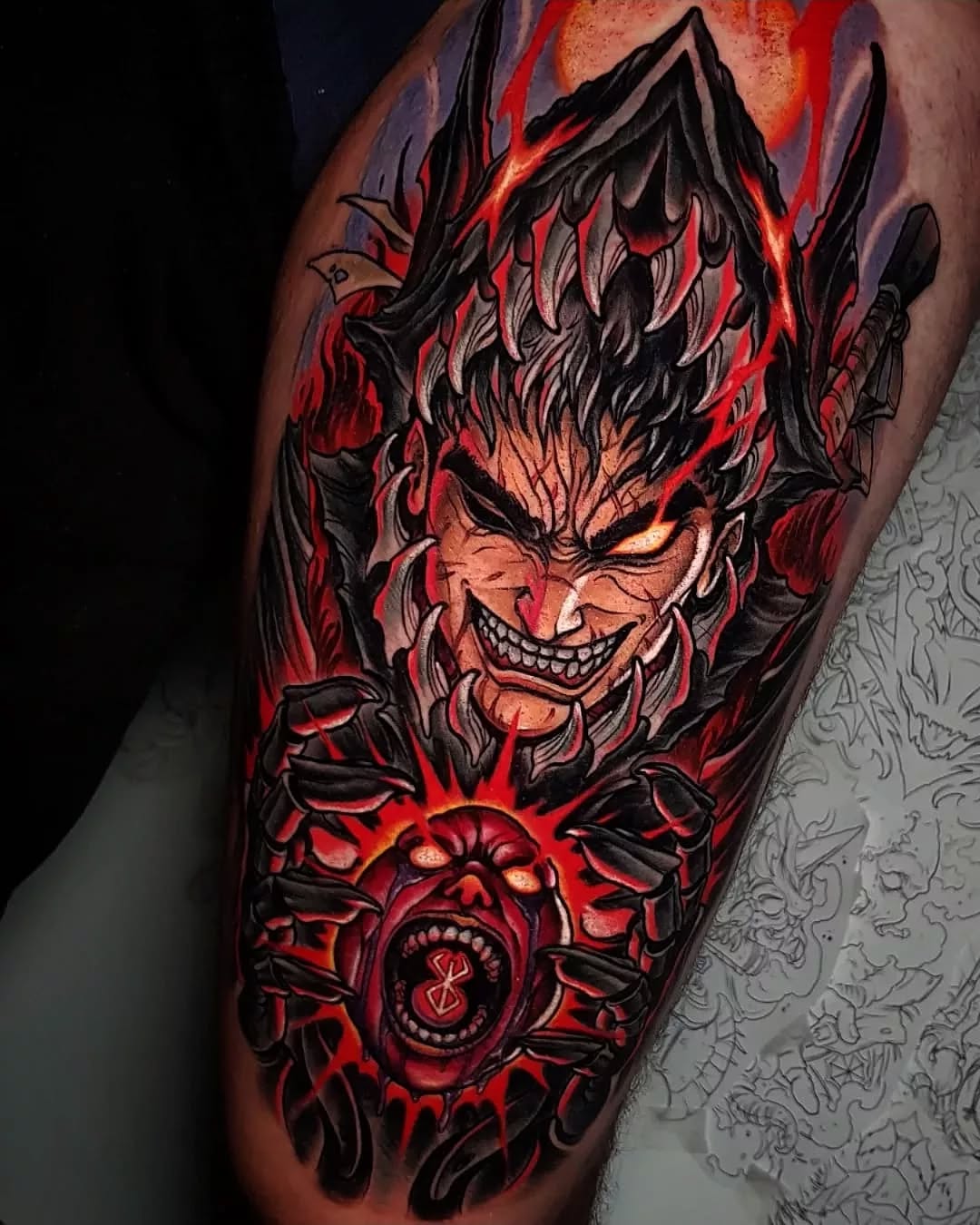 Demonic face tattoo with large horns, sharp teeth, and glowing red eyes surrounded by flames and a decorative circular symbol below, rendered in red and black on the upper arm.